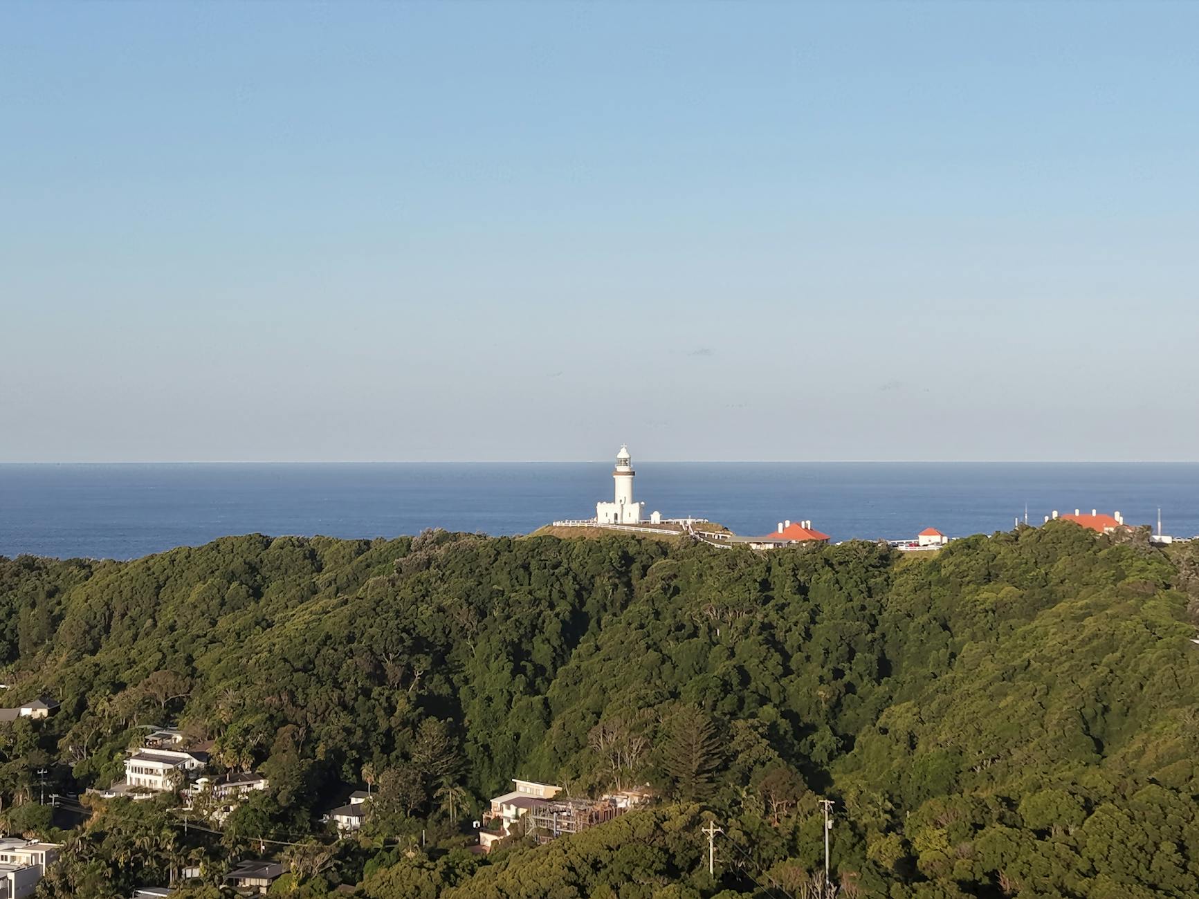 White lighthouse on a forested headland overlooking the blue ocean, with a few red-roofed buildings nearby.