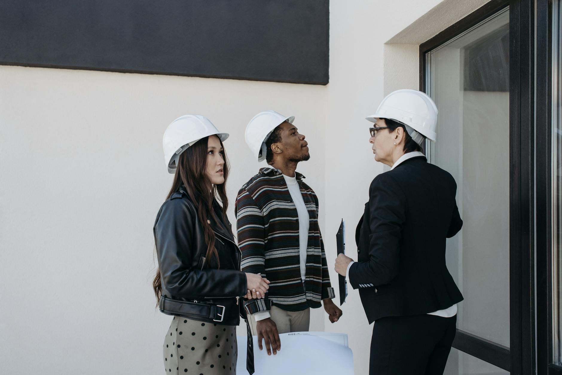 Three construction professionals in white hard hats discuss blueprints outside a building.