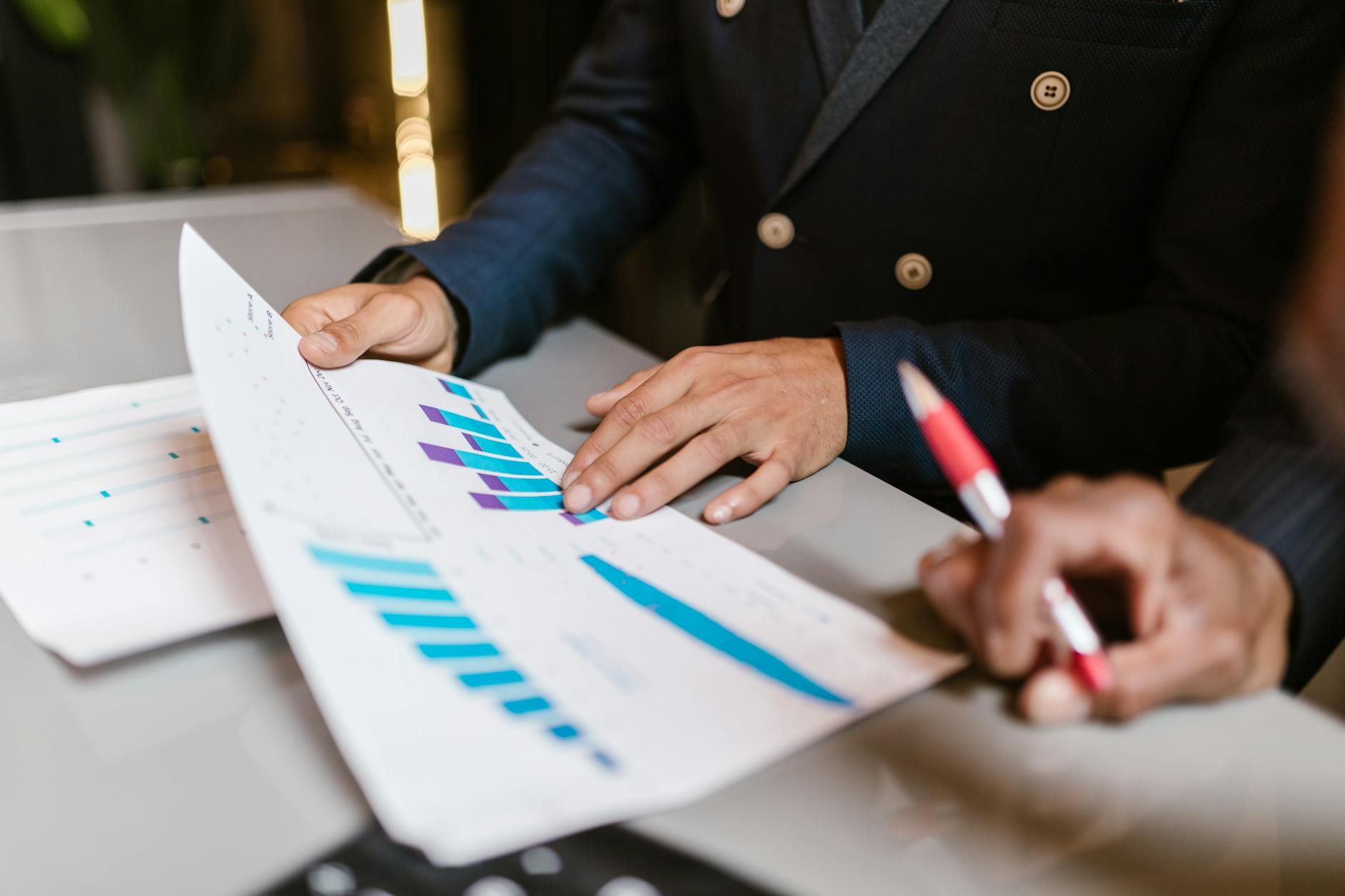 Two professionals review a printed report with blue bar charts at a desk, one pointing and the other writing with a red pen
