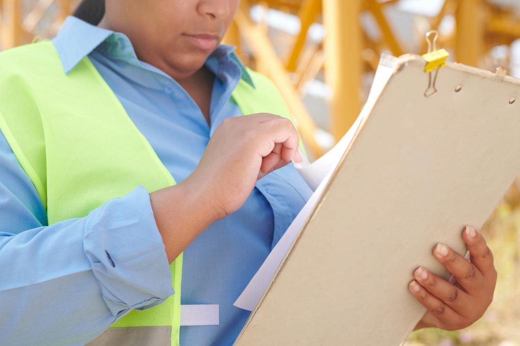 Construction worker in a neon safety vest and blue shirt writes on a clipboard outdoors at a construction site.
