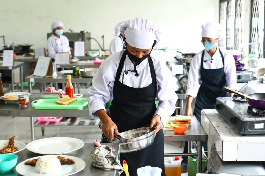 Chef in white uniform and mask mixing ingredients in a stainless steel bowl in a busy kitchen.
