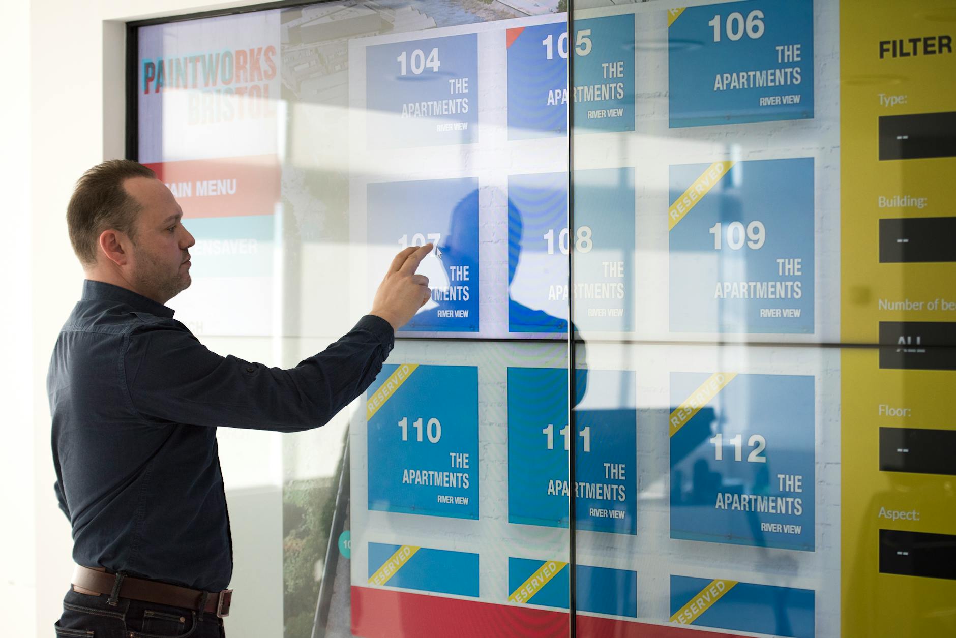 Man in a dark shirt touches a glass wall covered with blue apartment posters labeled 104–112 and 'The Apartments River View'.