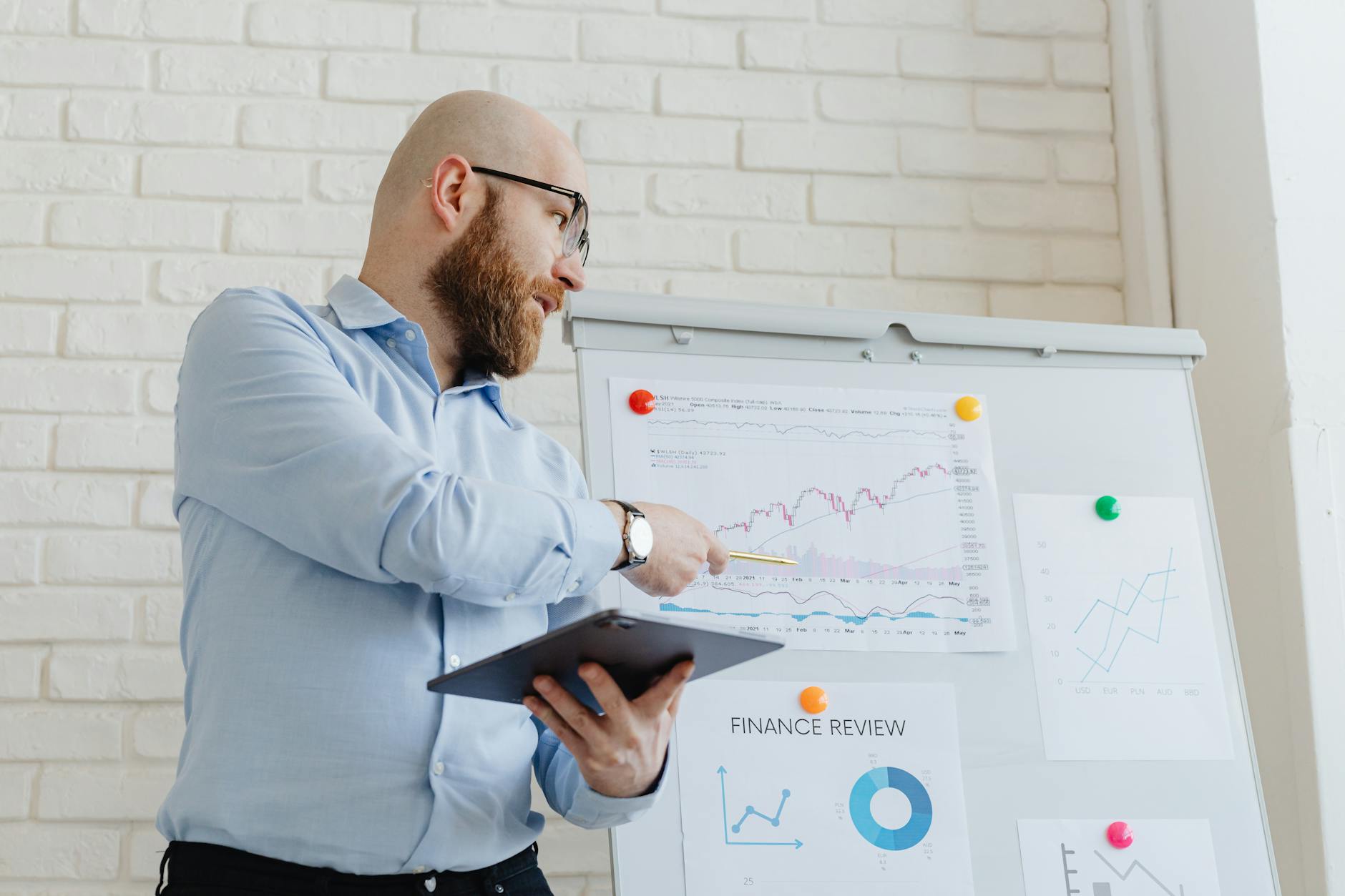 Bearded man in a light blue shirt points at financial charts on a whiteboard during a presentation.