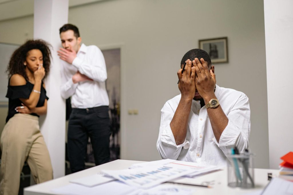 Man in a white shirt sits at a desk, hands covering his face; two coworkers stand in the blurred background in an office setting.