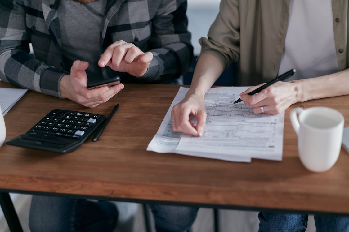 Janet and Kevin review documents at a wooden table, one annotating with a pen while the other uses a smartphone and calculator sits nearby
