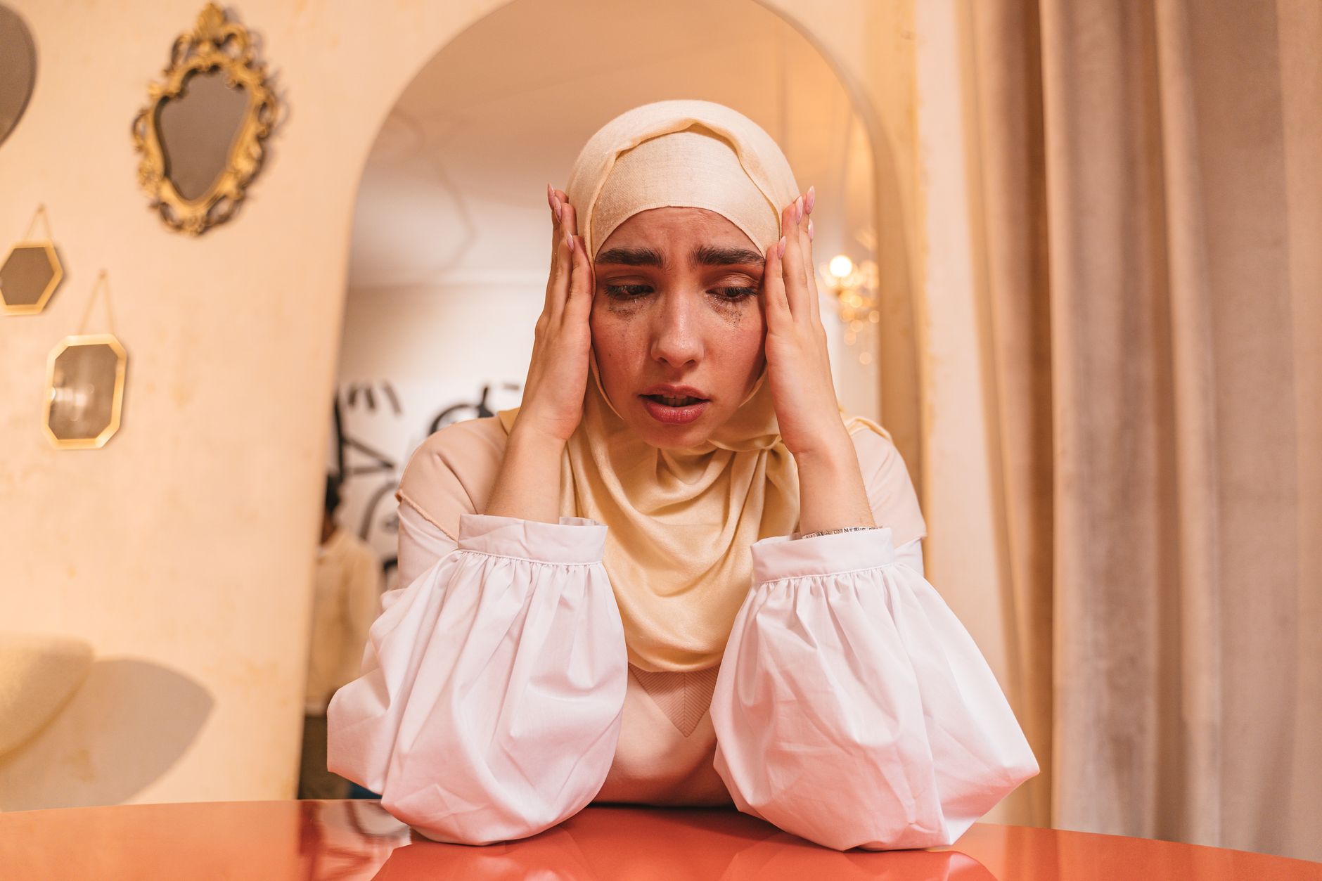 Woman in a beige hijab and pale pink top sits at an orange table, pressing her temples with both hands and looking down in distress.