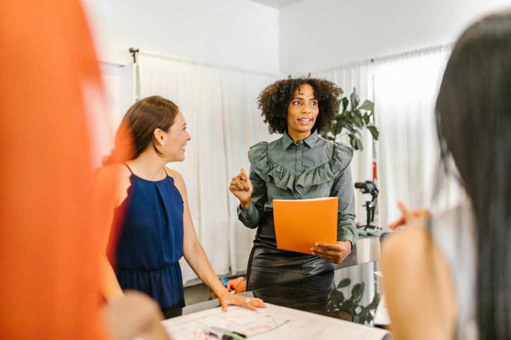 A diverse group of coworkers gathered around a counter as a woman with curly hair speaks, holding an orange folder.