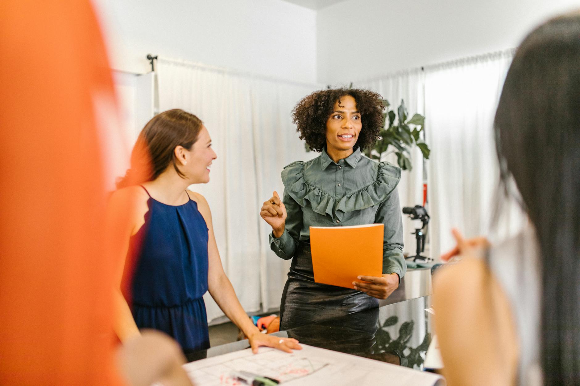 A diverse group of coworkers gathered around a counter as a woman with curly hair speaks, holding an orange folder.