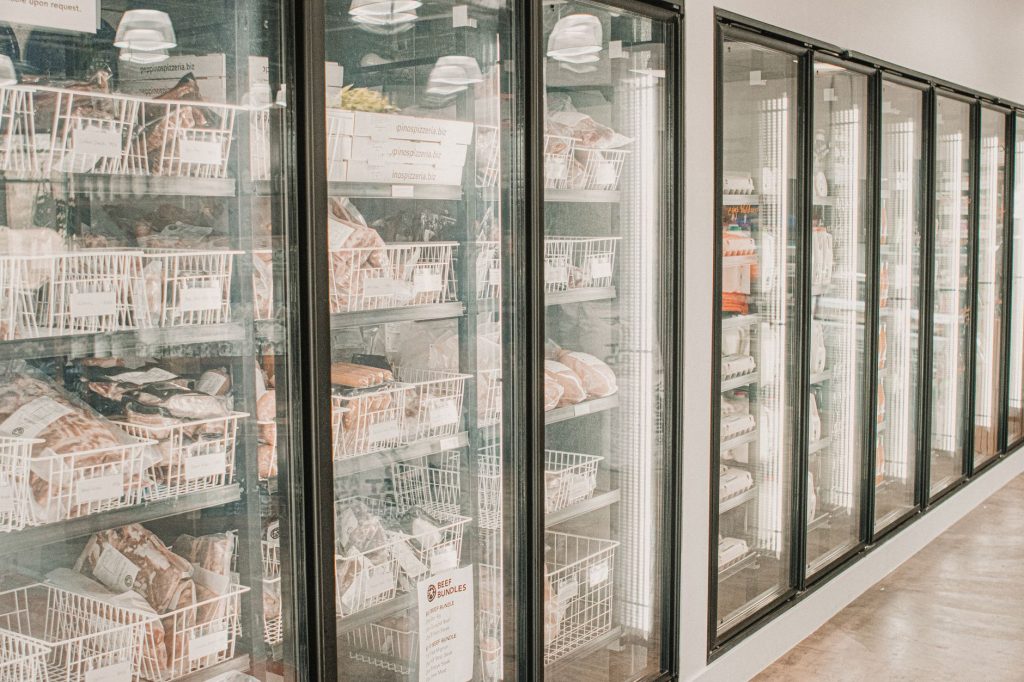Row of glass-door freezer cases in a grocery store, filled with packaged meat products in baskets and boxes behind the glass.