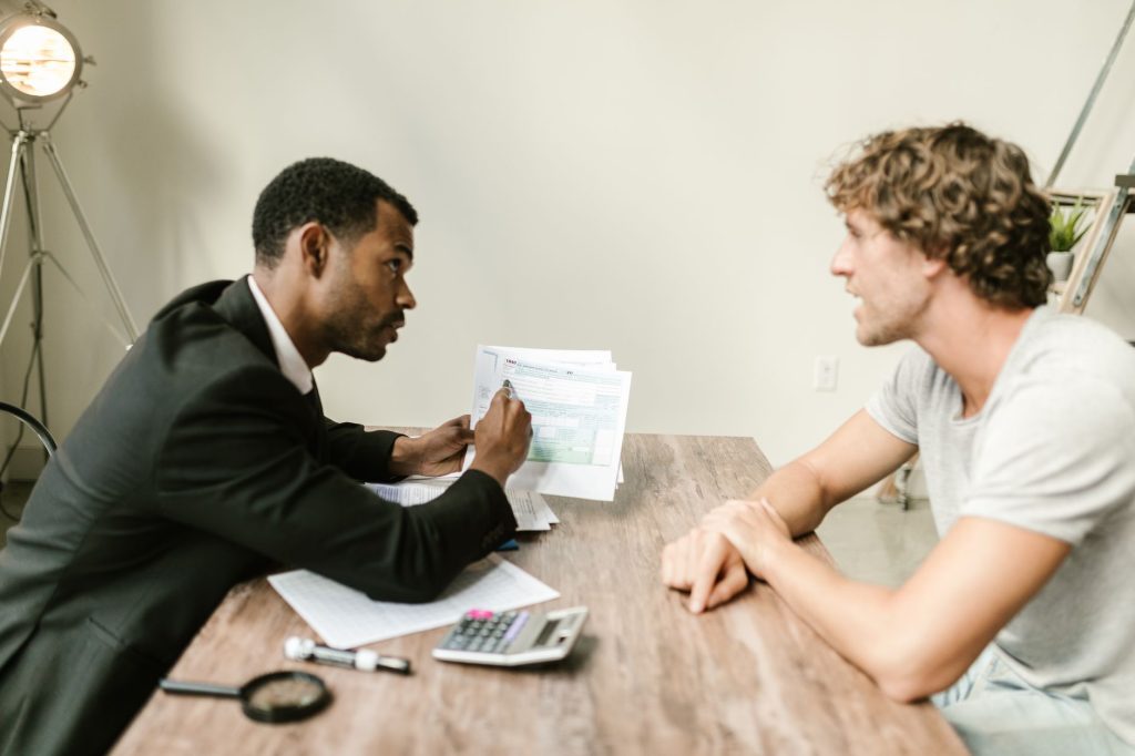 Two men sit at a wooden table reviewing documents, one in a suit pointing to papers as the other listens in a casual office setting.