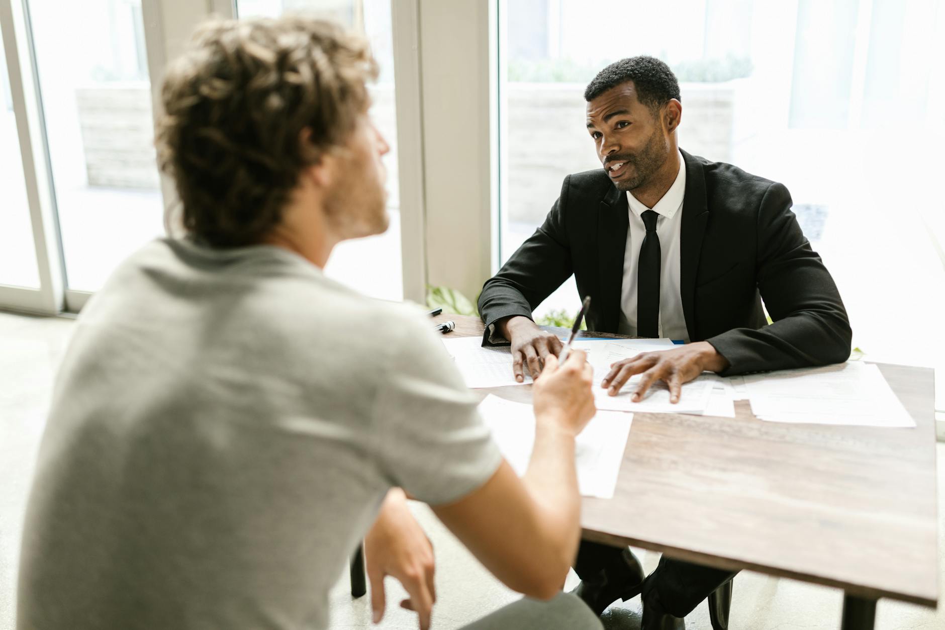 Two men sit at a table across from each other reviewing papers; one in a suit explains while the other listens.