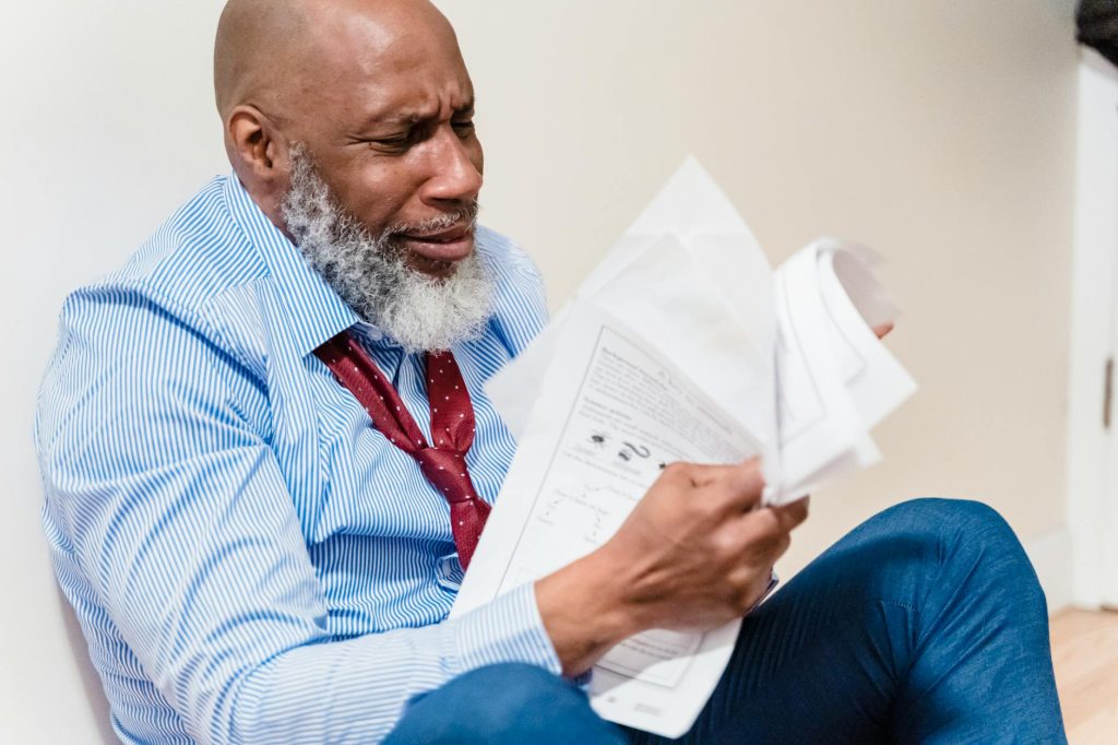 Bearded older man in a striped shirt and red tie sits against a wall, reading a stack of papers with a strained expression, showing concentration or frustration in a office-like setting.