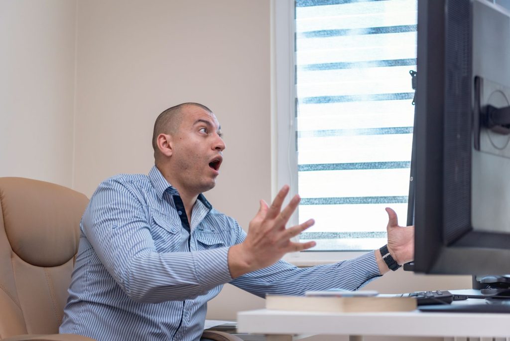 Man in a striped shirt sits at a desk, eyes wide, hands raised in surprise at a computer monitor in front of him.