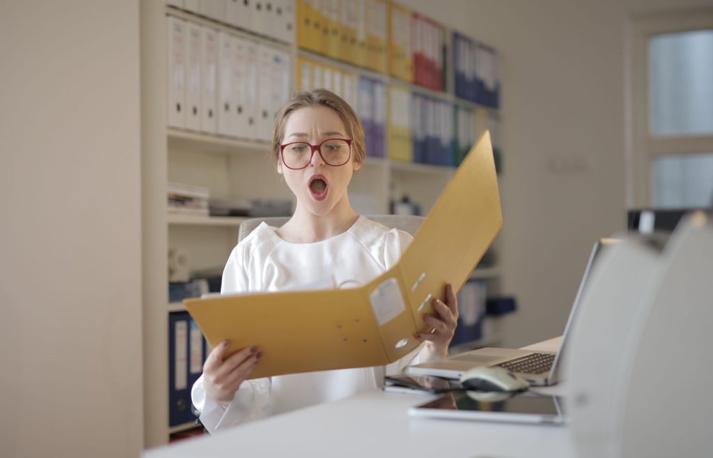 Woman in an office with red glasses opens a yellow file folder, looking surprised at a laptop on the desk in front of her.