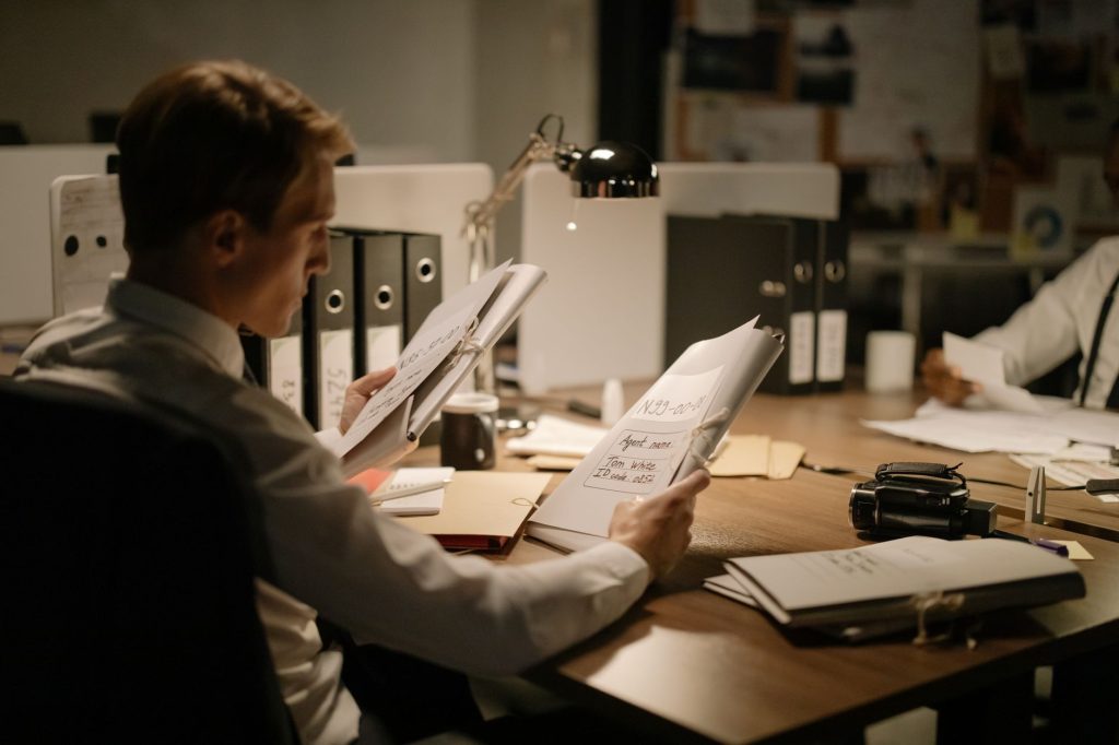 Two office workers review documents at a cluttered desk under a desk lamp.